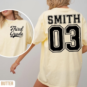 May include: Pale yellow t-shirt with "Third Grade" in black script on the front. The back displays "SMITH" above the number "03" in a black and white collegiate style. The shirt has a loose fit.