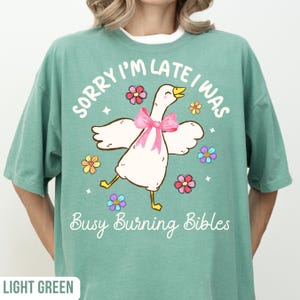May include: Light green t-shirt with the text "SORRY I'M LATE I WAS" above a cartoon goose wearing a pink bow and surrounded by colorful flowers. The text "Busy Burning Bibles" is below the goose.