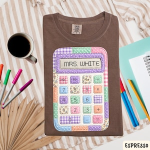 May include: Espresso-colored t-shirt featuring a colorful calculator design with the text "MRS. WHITE". The calculator design has a patchwork pattern with various colors and floral prints. The shirt is a Comfort Colors brand.