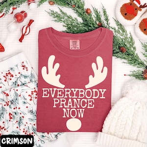 Peut inclure: T-shirt rouge cramoisi avec le texte "EVERYBODY PRANCE NOW" et des bois et un nez de renne cr&egrave;me. Le t-shirt fait partie d'un ensemble sur le th&egrave;me des f&ecirc;tes, comprenant un pantalon de pyjama &agrave; motifs et un bonnet en tricot blanc.