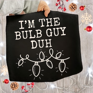 Peut inclure: Serviette de cuisine noire sur le th&egrave;me de No&euml;l avec le texte "I'M THE BULB GUY DUH" en blanc. La serviette pr&eacute;sente une guirlande d'ampoules blanches et est tenue par une personne. Des d&eacute;corations festives, dont des ornements rouges et des pommes de pin, entourent la serviette.