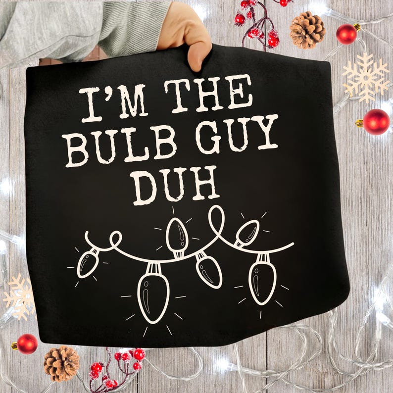 Peut inclure: Serviette de cuisine noire sur le th&egrave;me de No&euml;l avec le texte "I'M THE BULB GUY DUH" en blanc. La serviette pr&eacute;sente une guirlande d'ampoules blanches et est tenue par une personne. Des d&eacute;corations festives, dont des ornements rouges et des pommes de pin, entourent la serviette.