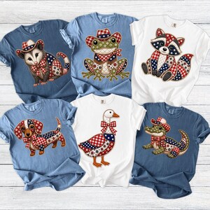 May include: Collection of t-shirts featuring patriotic animal designs. The shirts are in various colors, including blue and white. Each shirt showcases a different animal, such as a raccoon, frog, and dachshund, adorned with red, white, and blue patterns.