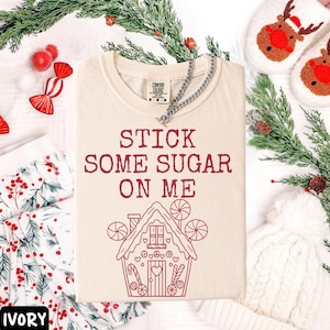 Peut inclure: T-shirt ivoire avec l'inscription "STICK SOME SUGAR ON ME" en bordeaux et un dessin de maison en pain d'&eacute;pices. Un collier en argent est pos&eacute; sur le t-shirt. D&eacute;corations de No&euml;l en arri&egrave;re-plan.