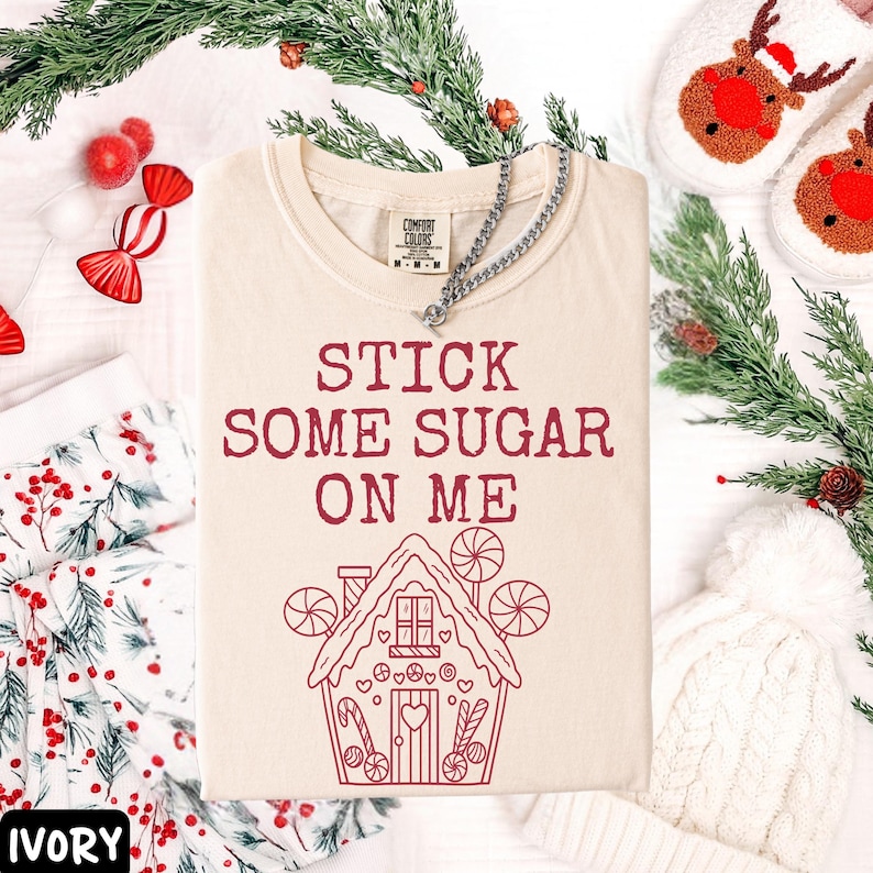 Peut inclure: T-shirt ivoire avec l'inscription "STICK SOME SUGAR ON ME" en bordeaux et un dessin de maison en pain d'&eacute;pices. Un collier en argent est pos&eacute; sur le t-shirt. D&eacute;corations de No&euml;l en arri&egrave;re-plan.