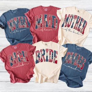 May include: A collection of six short-sleeved t-shirts in various colors, including blue, red, and cream. Each shirt features a different text design in a patriotic red, white, and blue pattern, with phrases like "Mother," "Maid," and "Bride."