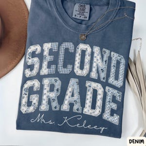 May include: Denim blue t-shirt with the words "SECOND GRADE" in a patchwork design with floral and gingham patterns. The name "Mrs. Keeley" is written below the text. A silver necklace is draped on the shirt.