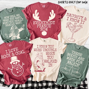 Peut inclure: Plusieurs t-shirts de couleurs vari&eacute;es, chacun orn&eacute; d'un motif et d'un texte sur le th&egrave;me des f&ecirc;tes. Les motifs incluent le P&egrave;re No&euml;l, des bois de rennes, un bonhomme de neige et Bigfoot. Le texte sur les chemises comprend des phrases comme "Naughty & I Know It".