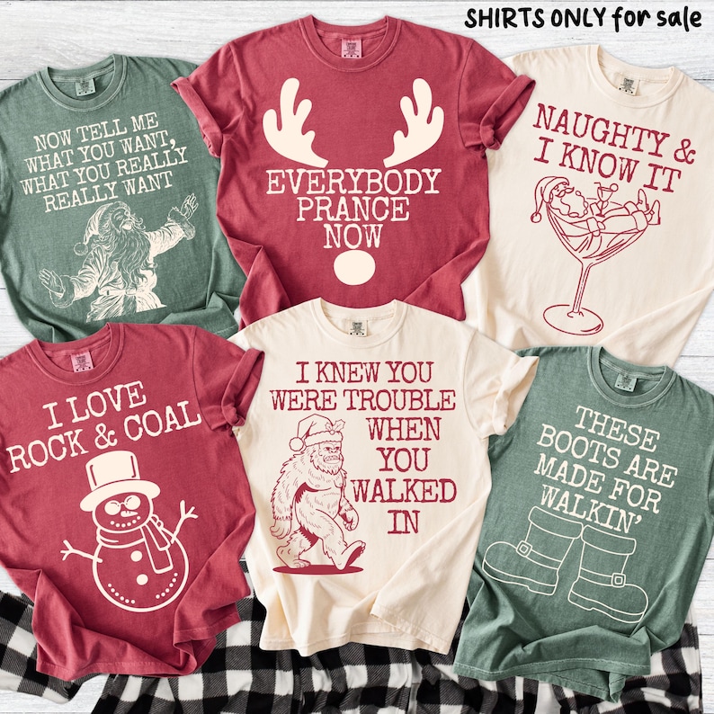 Peut inclure: Plusieurs t-shirts de couleurs vari&eacute;es, chacun orn&eacute; d'un motif et d'un texte sur le th&egrave;me des f&ecirc;tes. Les motifs incluent le P&egrave;re No&euml;l, des bois de rennes, un bonhomme de neige et Bigfoot. Le texte sur les chemises comprend des phrases comme "Naughty & I Know It".