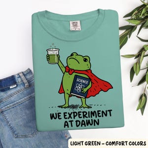 May include: A light green t-shirt featuring a graphic of a green frog wearing a red cape, holding a beaker of green liquid and a blue book labeled SCIENCE. Below the frog, the text reads WE EXPERIMENT AT DAWN in black capital letters.