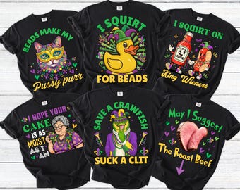 May include: Black t-shirts with colorful Mardi Gras-themed graphics. Designs include a cat with a mask, a duck with a jester hat, a bottle of hot sauce, a hot dog, a woman with a cake, a frog, and a roast beef.