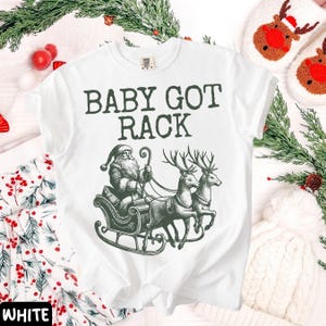 Peut inclure: T-shirt blanc avec l'inscription "BABY GOT RACK" en vert fonc&eacute;. Le motif repr&eacute;sente le P&egrave;re No&euml;l dans un tra&icirc;neau tir&eacute; par deux rennes. Un design festif pour les f&ecirc;tes.