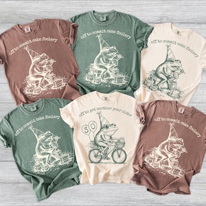 May include: A collection of t-shirts in various colors, each featuring a whimsical frog graphic. The frog is depicted with a party hat, surrounded by cakes, and the text "off to commit cake foolery" or "off to get another year older" is printed above the frog.