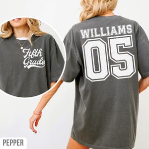 May include: A dark grey t-shirt with the words "Fifth Grade" in white on the front. The back features the name "WILLIAMS" and the number "05" in a white, outlined font. The shirt has a loose fit.