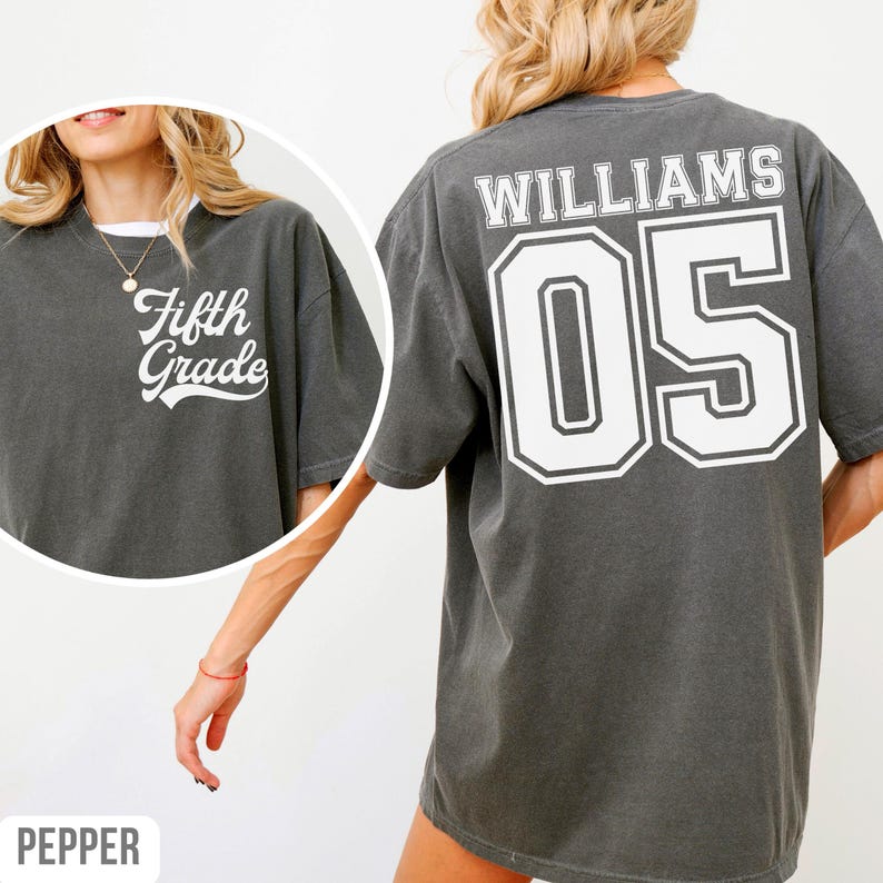 May include: A dark grey t-shirt with the words "Fifth Grade" in white on the front. The back features the name "WILLIAMS" and the number "05" in a white, outlined font. The shirt has a loose fit.