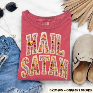 May include: Crimson-colored t-shirt with the words "HAIL SATAN" embroidered in floral letters. The shirt is folded and surrounded by accessories, including sunglasses, a necklace, denim shorts, and sandals. The text "CRIMSON - COMFORT COLORS" is at the bottom.