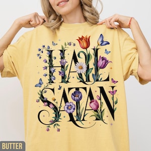 May include: Yellow t-shirt with the words "HAIL SATAN" in black, decorated with floral and butterfly graphics. The design includes purple, red, and white flowers, and blue and pink butterflies. The shirt is a casual, comfortable style.