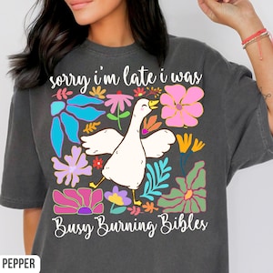 May include: Dark gray t-shirt with a whimsical design featuring a white goose with outstretched wings, surrounded by colorful flowers. The text "sorry i'm late i was Busy Burning Bibles" is printed above and below the goose.