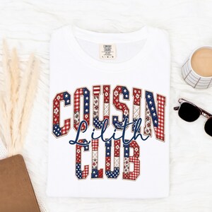 May include: White t-shirt with the text "COUSIN Lilith CLUB" in red, white, and blue patriotic colors. The design features stars and plaid patterns. A coffee cup and sunglasses are visible in the background.