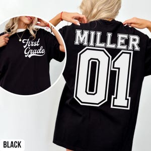 May include: Black t-shirt with white text. The front says "First Grade" in a cursive font. The back of the shirt features the name "MILLER" above the number "01" in a white, block font. The word "BLACK" is at the bottom.