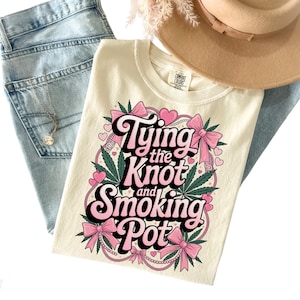 May include: Ivory-colored t-shirt with the text "Tying the Knot and Smoking Pot" in a pink and black design. The design includes pink bows, hearts, and cannabis leaves. The shirt is laid on top of blue jeans and a tan hat.