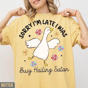 May include: Yellow t-shirt with the text "SORRY I'M LATE I WAS Busy Hailing Satan" and a cartoon goose illustration. The goose is white with yellow feet and a yellow beak, surrounded by colorful flowers.