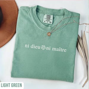 May include: Light green Comfort Colors t-shirt with the text "ni dieu ni maître" in white. A silver necklace with a pendant rests on the shirt. The shirt is a size small.