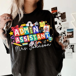 May include: Black t-shirt with a colorful graphic that reads "ADMIN ASSISTANT" in large, block letters. The design includes a rainbow, pencil, and other school-related icons. The name "Mrs. Johnson" is written below the text.