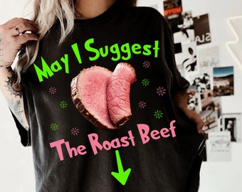 May include: Black t-shirt with the text "May I Suggest The Roast Beef" in green and pink. A graphic of a roast beef cut is in the center, with green and pink snowflake accents and a green arrow pointing down.