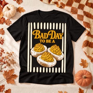 May include: A black short-sleeve t-shirt featuring a graphic print with the text "BAD DAY TO BE A" in yellow letters above three illustrations of deviled eggs, all framed by vertical white stripes on the sides of the design.