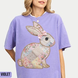 May include: A purple t-shirt with a patchwork bunny applique. The bunny is made of different fabrics in shades of pink, white, and brown. The bunny has a pink bow on its head.