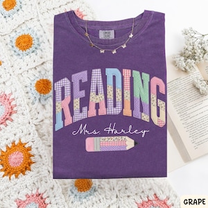 May include: A purple t-shirt featuring the word READING in a multi-colored patchwork pattern, the text Mrs. Harley in white script, and a pencil graphic with a patchwork design. A gold butterfly necklace rests on the collar of the shirt.