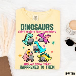 May include: A pale yellow t-shirt featuring a graphic of five colorful dinosaurs using computers. The text reads: DINOSAURS didn't update their computers and we know what HAPPPENED TO THEM. The shirt has a crew neckline and a Comfort Colors brand label.