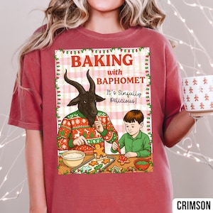 May include: Crimson-colored t-shirt featuring a humorous illustration of a goat-headed figure and a child baking gingerbread cookies. The text on the shirt reads "Baking with Baphomet. It's Sinfully Delicious!" The design is framed with festive lights.
