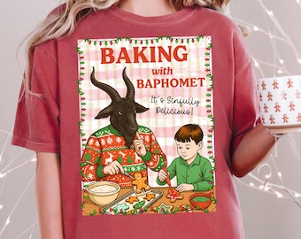May include: Crimson-colored t-shirt featuring a humorous illustration of a goat-headed figure and a child baking gingerbread cookies. The text on the shirt reads "Baking with Baphomet. It's Sinfully Delicious!" The design is framed with festive lights.