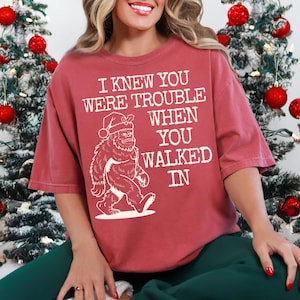 Peut inclure: Un t-shirt rouge-brun avec le texte "I KNEW YOU WERE TROUBLE WHEN YOU WALKED IN" et un dessin de Bigfoot portant un bonnet de P&egrave;re No&euml;l. Le t-shirt est &agrave; manches courtes et de style d&eacute;contract&eacute;.