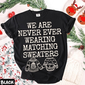 Peut inclure: T-shirt noir avec l'inscription blanche "WE ARE NEVER EVER WEARING MATCHING SWEATERS". Le t-shirt pr&eacute;sente des illustrations de deux pulls. Le mot "BLACK" est en bas &agrave; gauche de l'image. Th&egrave;me des f&ecirc;tes.