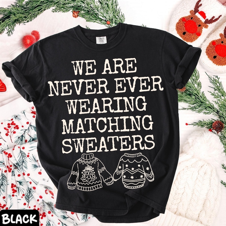 Peut inclure: T-shirt noir avec l'inscription blanche "WE ARE NEVER EVER WEARING MATCHING SWEATERS". Le t-shirt pr&eacute;sente des illustrations de deux pulls. Le mot "BLACK" est en bas &agrave; gauche de l'image. Th&egrave;me des f&ecirc;tes.