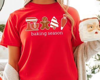 May include: A red t-shirt with a white graphic that says "baking season". The graphic includes a mixer, gingerbread man, Christmas tree, and pastry bag. The shirt is made of a soft material.