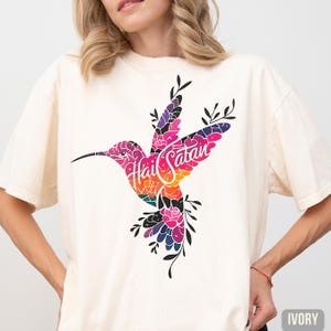May include: Ivory-colored t-shirt featuring a colorful hummingbird design. The hummingbird is filled with floral patterns in shades of pink, purple, orange, and green. The words "Hail Satan" are written across the hummingbird's body.