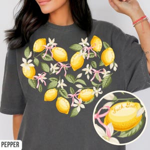May include: Dark gray t-shirt featuring a heart-shaped design of yellow lemons, white flowers, and green leaves. Each lemon has the text "Fuck God" on it. Pink ribbons accent the design. The word "PEPPER" is at the bottom left.