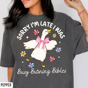 May include: Dark gray t-shirt with a whimsical graphic of a white goose wearing a pink bow, surrounded by colorful flowers. The text reads "SORRY I'M LATE I WAS Busy Burning Bibles."