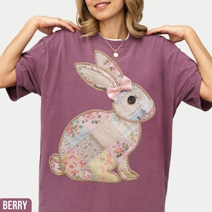 May include: A purple t-shirt with a patchwork bunny applique. The bunny is made of different fabrics in shades of pink, white, and yellow. The bunny has a pink bow on its head.
