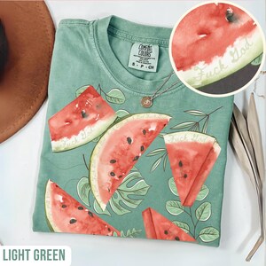 May include: Light green t-shirt with a watermelon design. The shirt features watercolor-style watermelon slices with black seeds and the text "Fuck God". The shirt has a crew neck and short sleeves. A silver necklace with a pendant is visible.