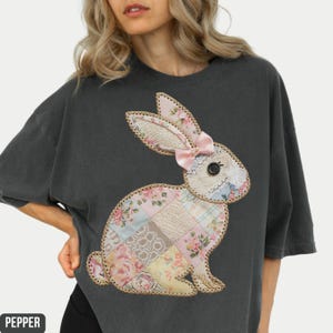 May include: A dark gray t-shirt with a patchwork bunny applique. The bunny is made of various fabrics in shades of pink, white, and blue, with floral patterns. The bunny has a pink bow on its head.