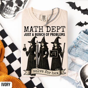 May include: Ivory-colored t-shirt with the text "MATH DEPT JUST A BUNCH OF PROBLEMS" and an illustration of three witches. The witches are holding a flask, a pencil, and a sign with the square root of -1. The phrase "solve for hex" is on a banner.