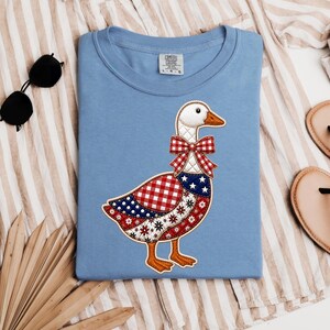 May include: A light blue Comfort Colors t-shirt featuring a patriotic goose design. The goose has a white head, orange beak, and a red and white checkered bow. Its body is a patchwork of red, white, and blue patterns.