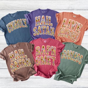 May include: Six short-sleeved t-shirts in various colors, including blue, purple, and pink. Each shirt features a different phrase in large, floral-filled letters: "UNHOLY", "HAIL SATAN", "LIVE DELICIOUSLY", "BAPHOMET", and "GODLESS".