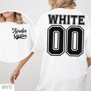 May include: White t-shirt with "Kinder Garten" in a cursive font on the front. The back displays "WHITE" above the number "00" in black and white. The shirt has a relaxed, oversized fit. A casual garment for everyday wear.