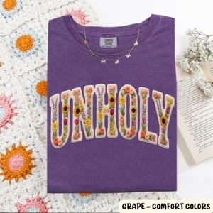 May include: A grape-colored t-shirt with the word "UNHOLY" embroidered with colorful flowers. A gold butterfly necklace rests on the shirt. The shirt is from Comfort Colors.